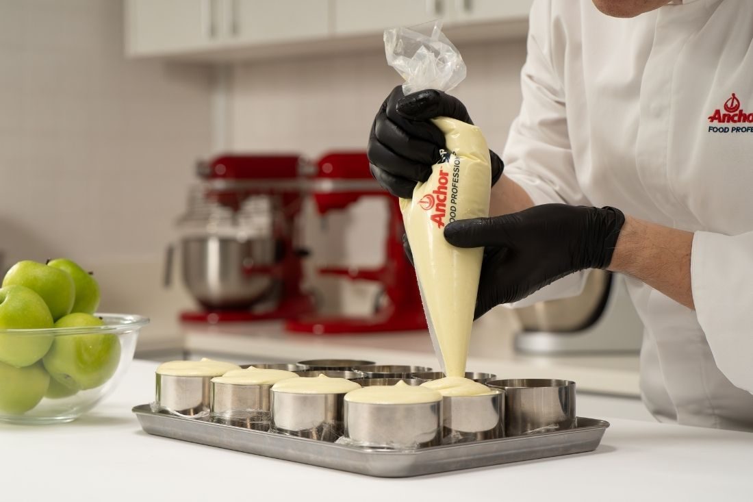 Pastry chef piping creamy dessert mixture into stainless‑steel molds in a professional kitchen.